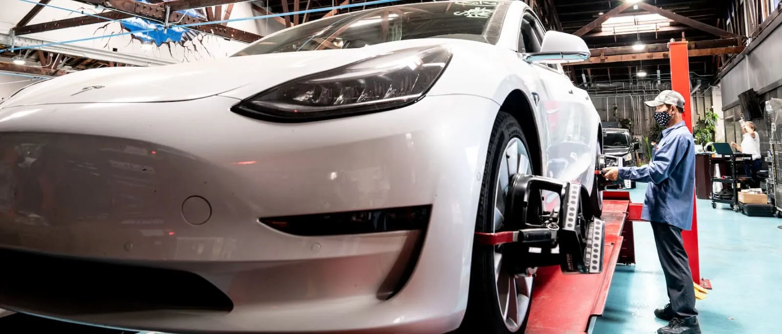 Technician with face mask calibrating the wheel alignment of a white Tesla car in an automotive workshop.