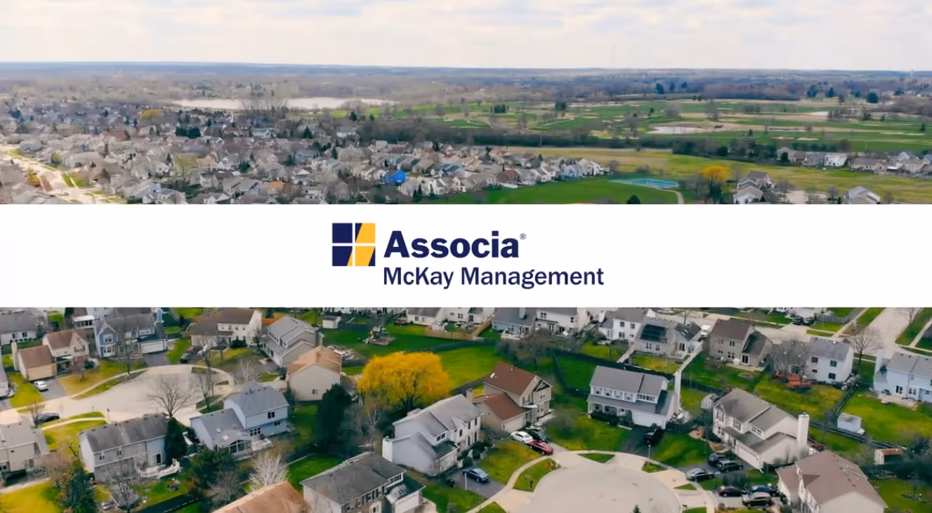 Aerial view of a suburban neighborhood with houses and green lawns under a partly cloudy sky, with Associa McKay Management logo overlayed in the center.