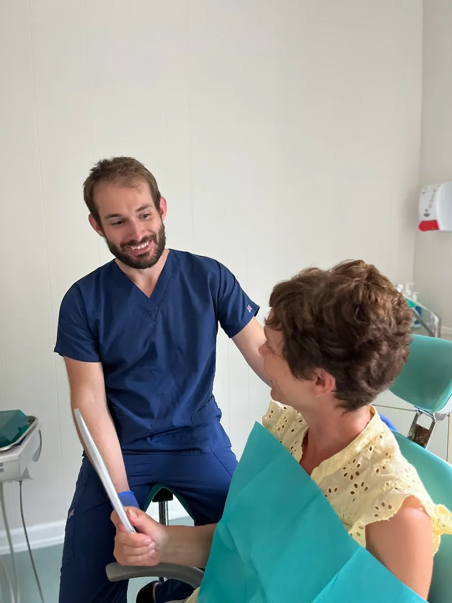 Dentist in blue scrubs smiling at a female patient holding a dental mirror in a clinic.