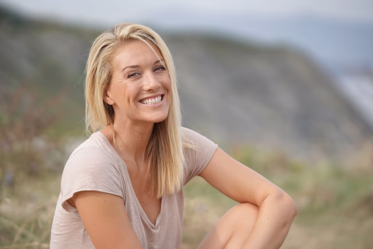Smiling woman with blonde hair sitting outdoors with a blurred natural background.