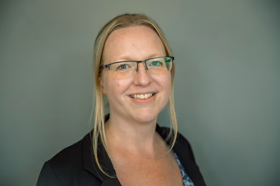 Smiling woman with glasses and blonde hair wearing a black blazer against a plain gray background.