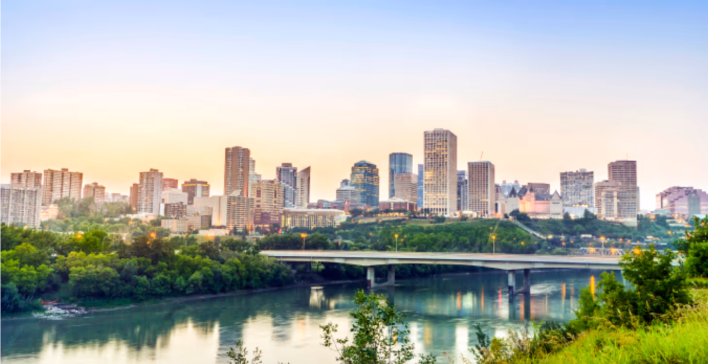Scenic view of Vancouver’s downtown skyline at sunrise with a bridge spanning over the water and lush green trees in the foreground.