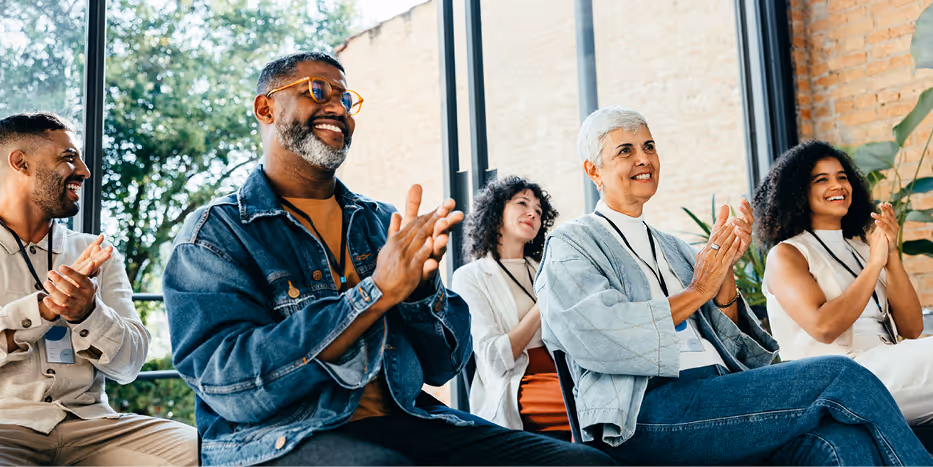 Group of smiling people of various ages and backgrounds sitting in chairs and applauding at an indoor event with large windows and natural light.