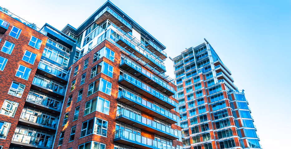 Upward view of two contemporary high-rise apartment buildings with red brick and large glass balconies under a clear blue sky.