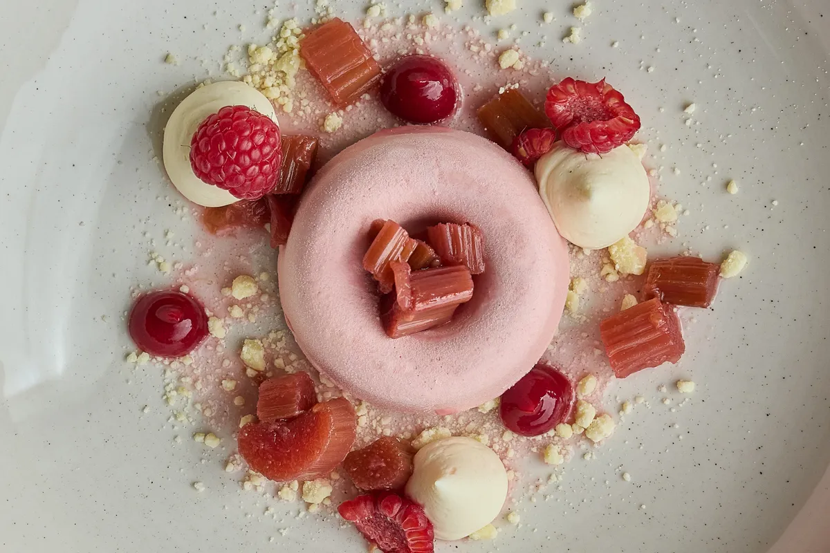 Rosa ringformad dessert med röda bär, små vit kräm och bitar av rabarber på en vit tallrik.