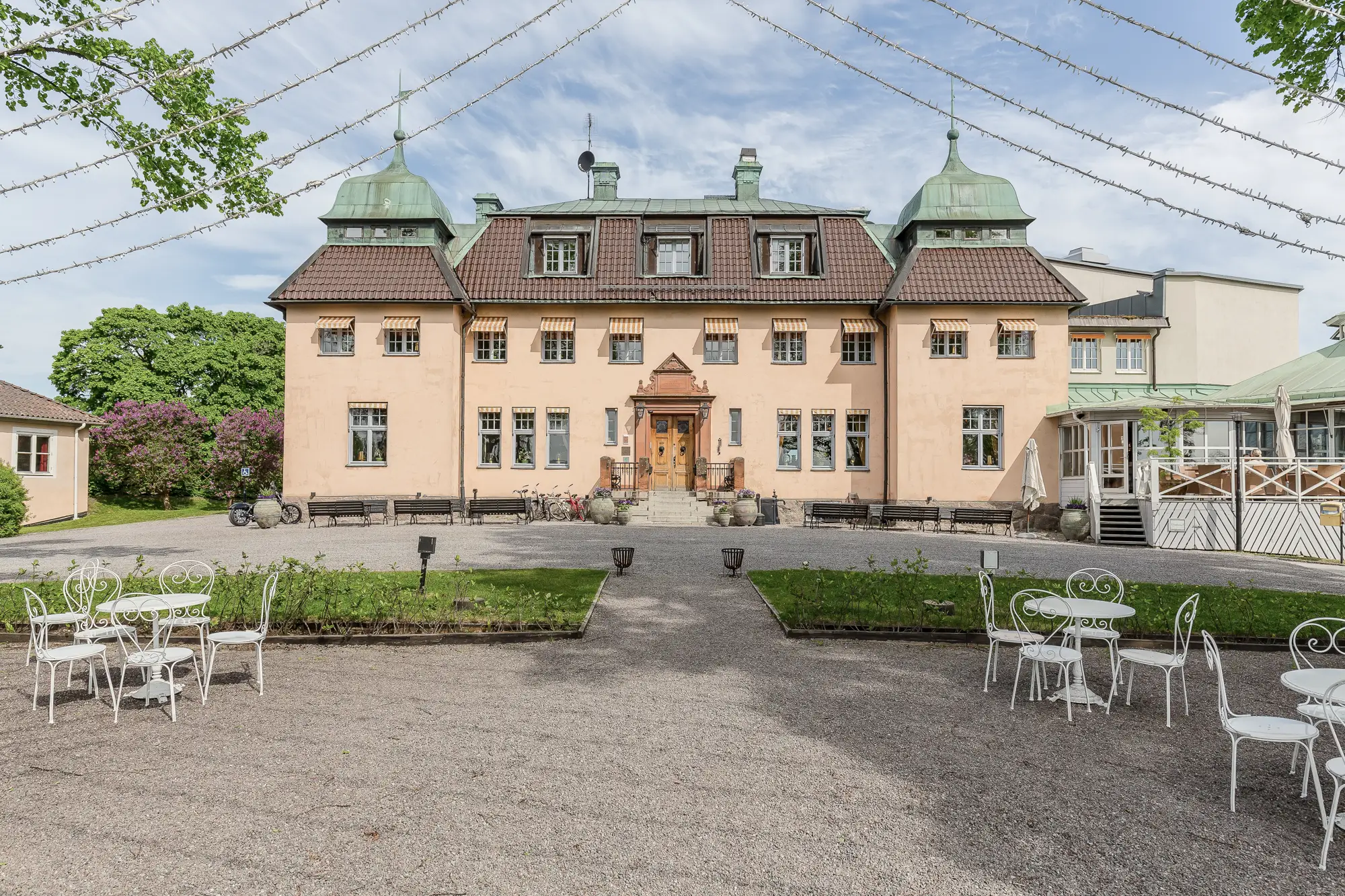 Ståtligt hus med gröna takkupoler och beige fasad omgiven av trädgård med utebord och stolar.