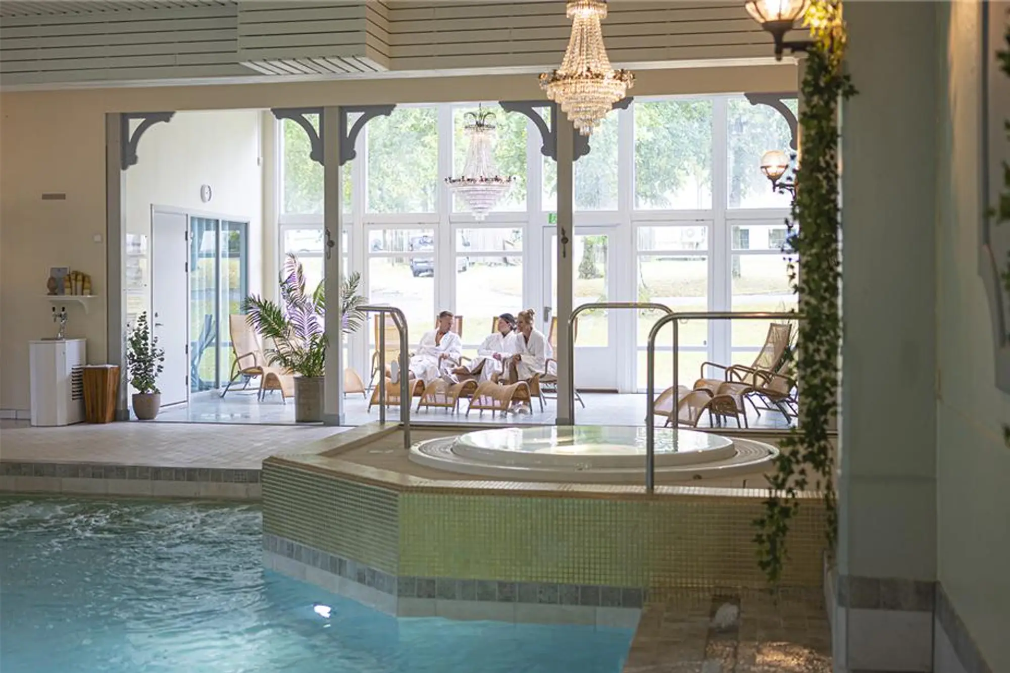 Personer i badrockar som sitter och kopplar av på korgstolar i ett ljust spa-område med pool och jacuzzi.