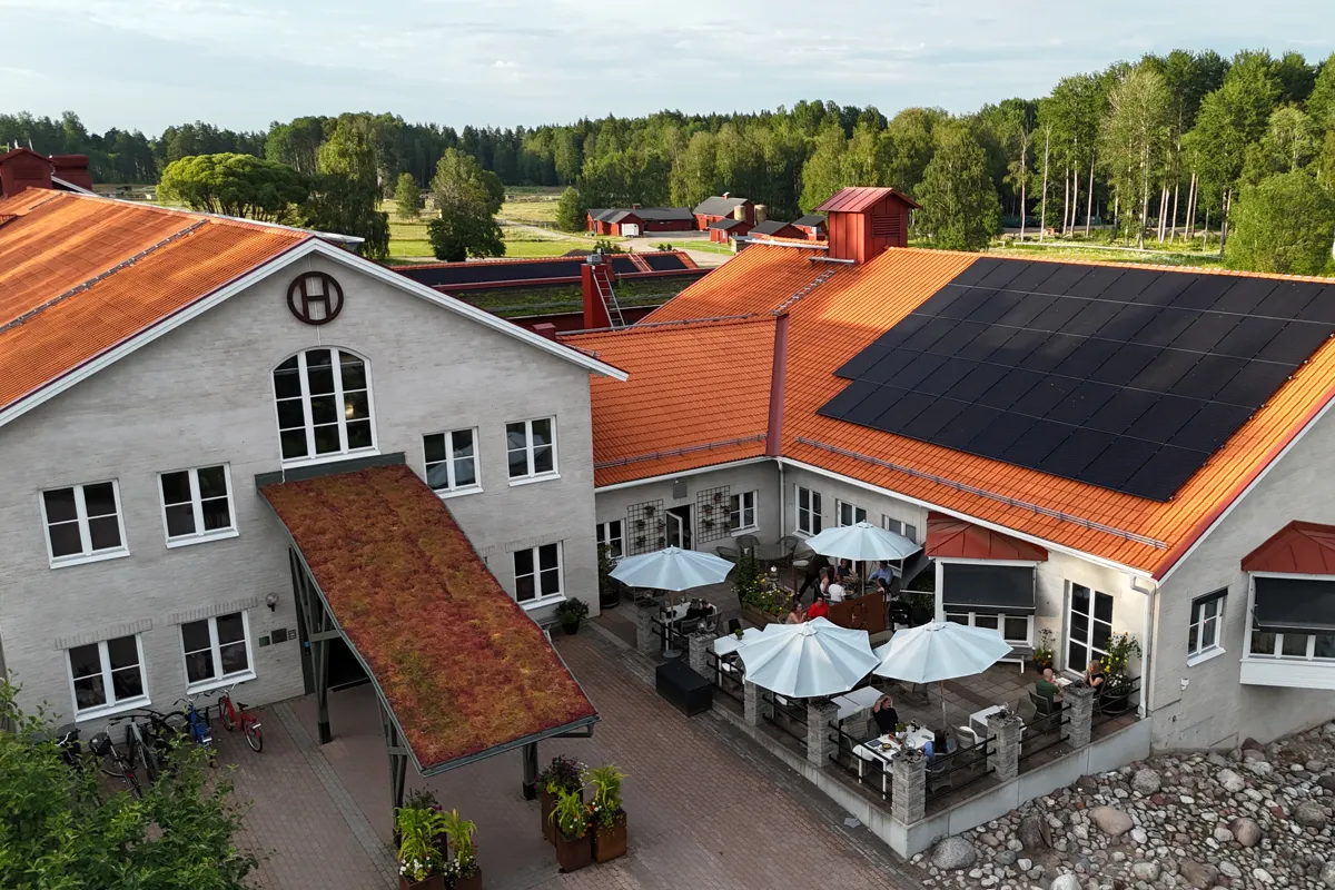 Flygfoto av en ljus byggnad med orange tegeltak och solpaneler, med en innergård där folk sitter under vita parasoller.