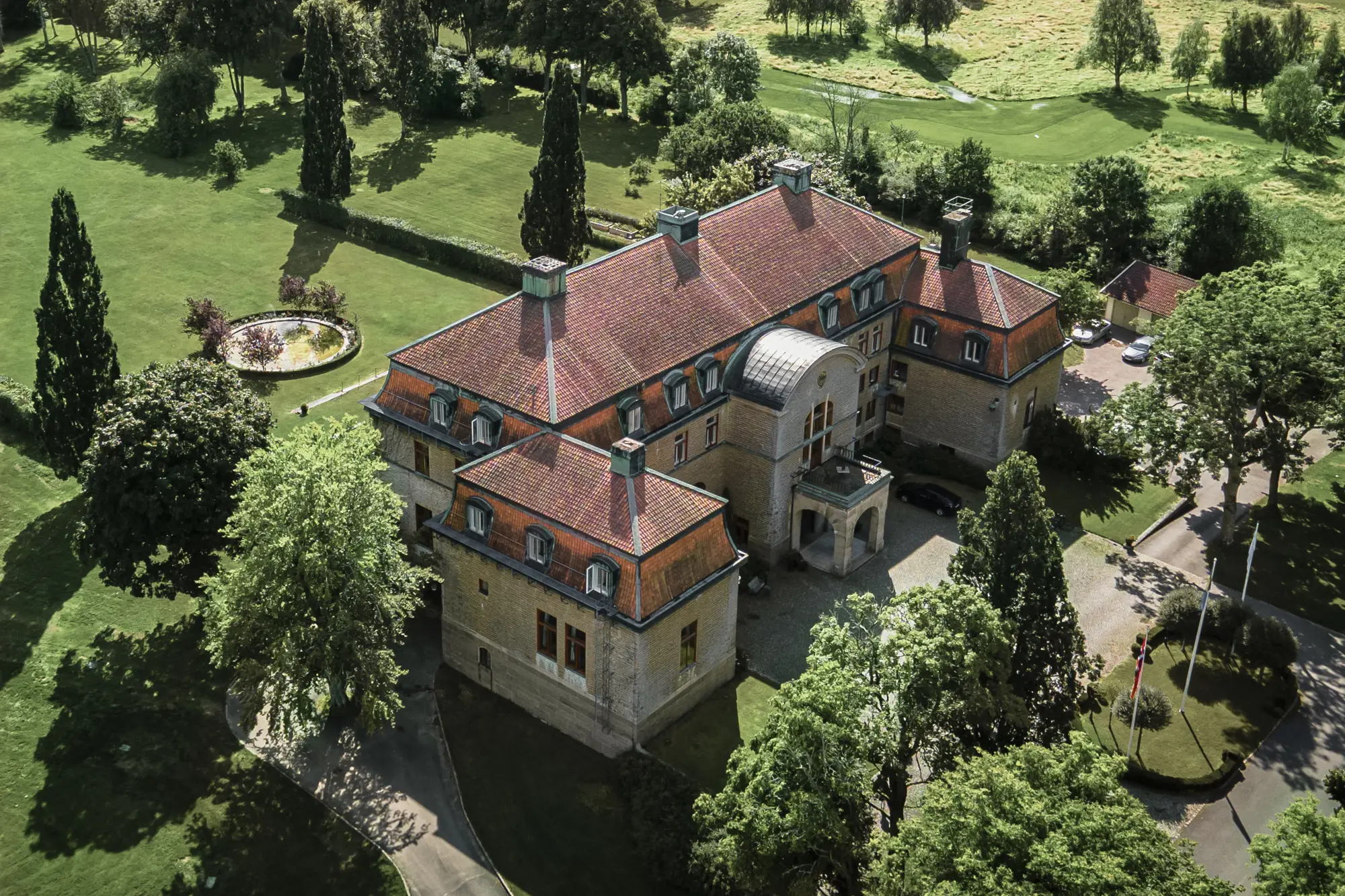 Flygbild på stort slott med rött tegeltak omgivet av grönskande trädgård och gräsmatta i sommarljus.