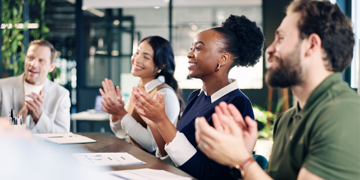 Colleagues applaud and smile during a meeting celebrating professional achievements and industry recognition.