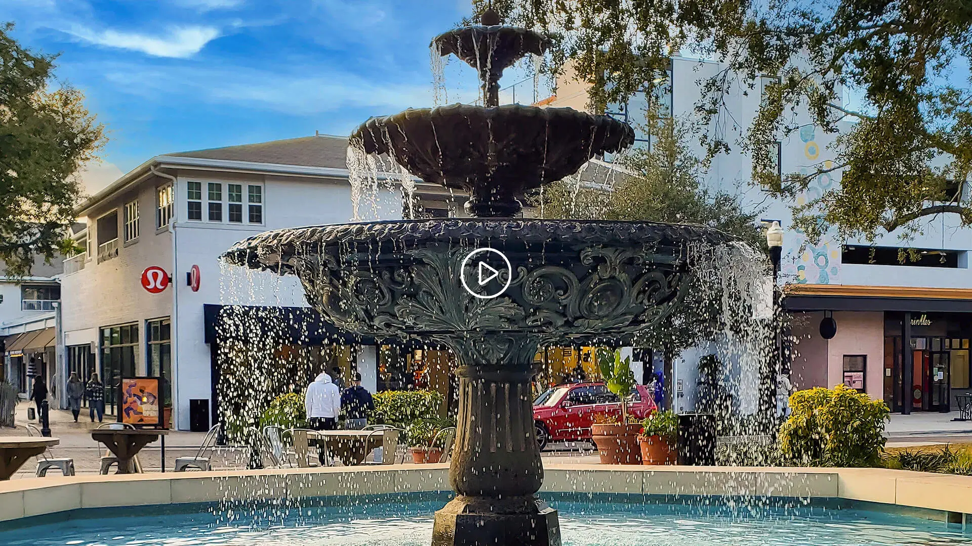 Tiered fountain with flowing water in a lively shopping district with with play button overlay.