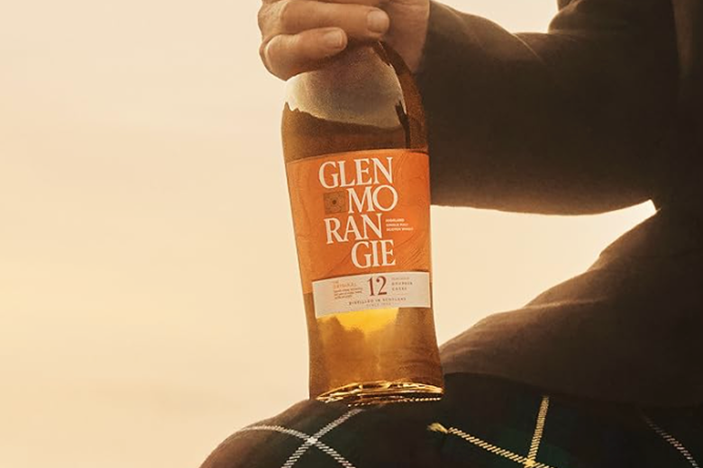 Hand holding a bottle of Glenmorangie 12-year-old single malt Scotch whisky resting on a tartan-patterned fabric.
