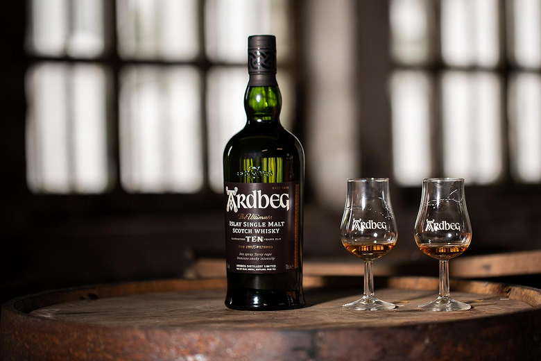 Bottle of Ardbeg 10-year-old Islay single malt Scotch whisky next to two partially filled Ardbeg branded whisky glasses on a wooden barrel.
