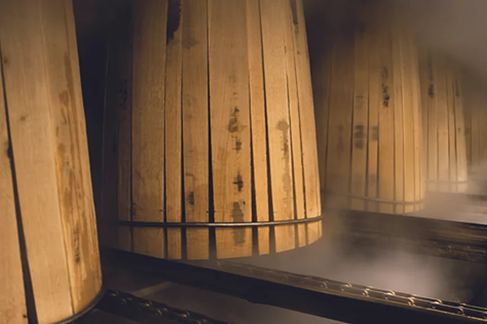 Rows of large wooden barrels with metal bands in a dimly lit, smoky environment, likely a storage or aging room.