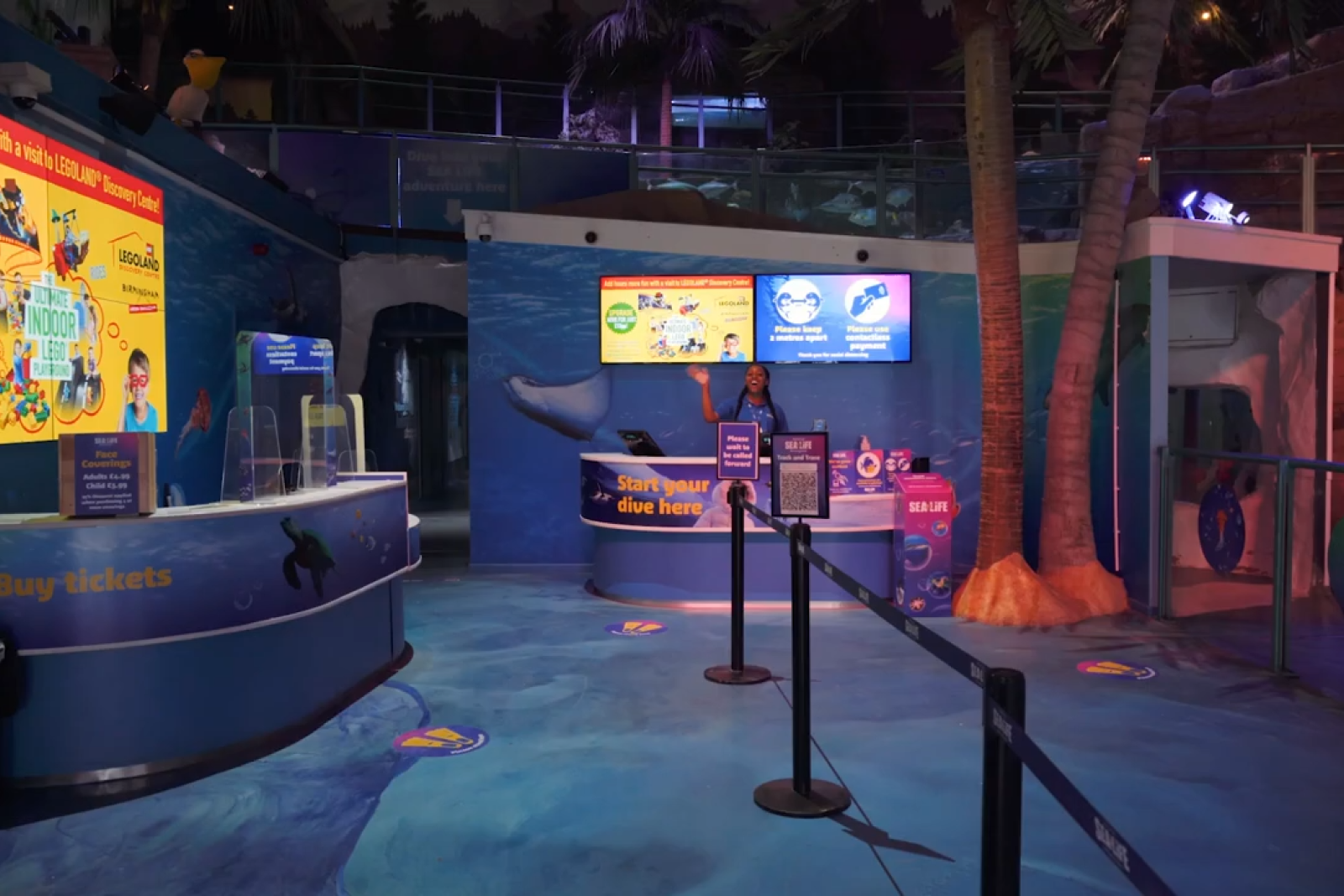 Reception area of an aquarium attraction with a staff member waving behind a counter that says 'Start your dive here,' underwater-themed decor, and digital screens displaying visitor information.