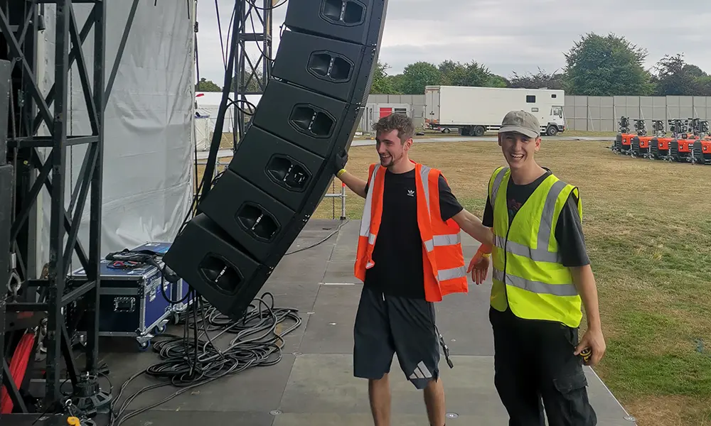 Two dBs students stand next to a flying speak stack in a festival