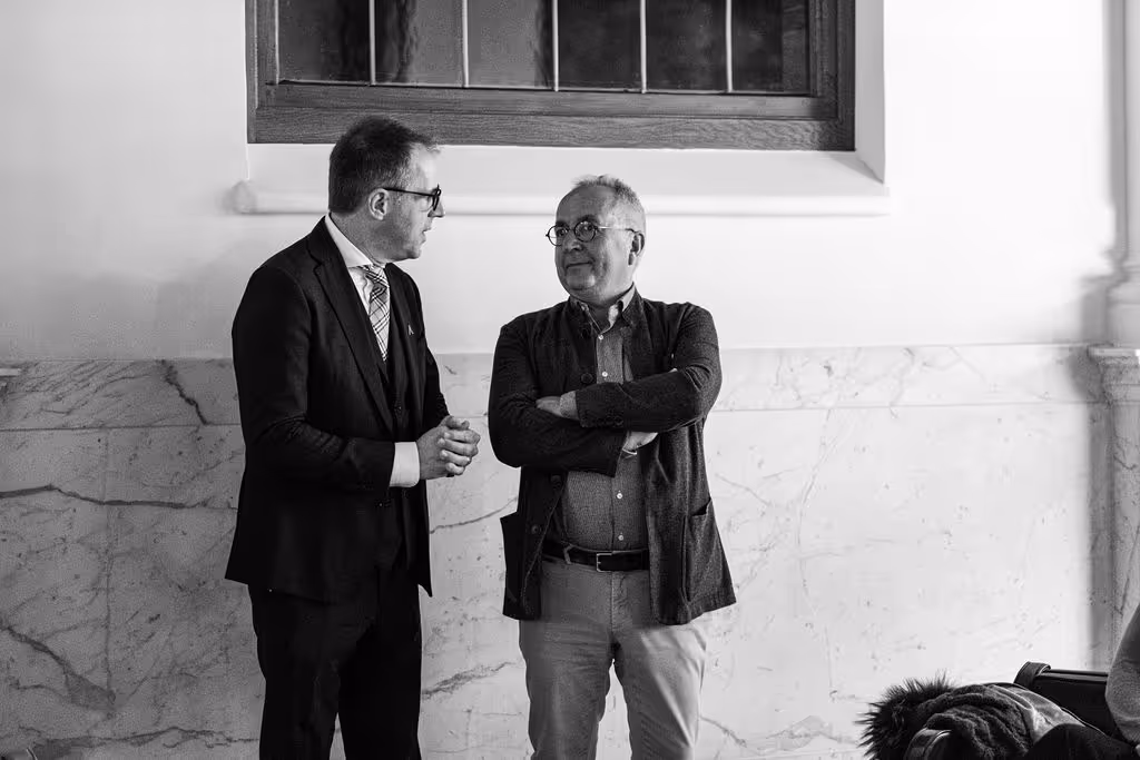 Two smartly-dressed men talking at the side of room with marble detail on wall