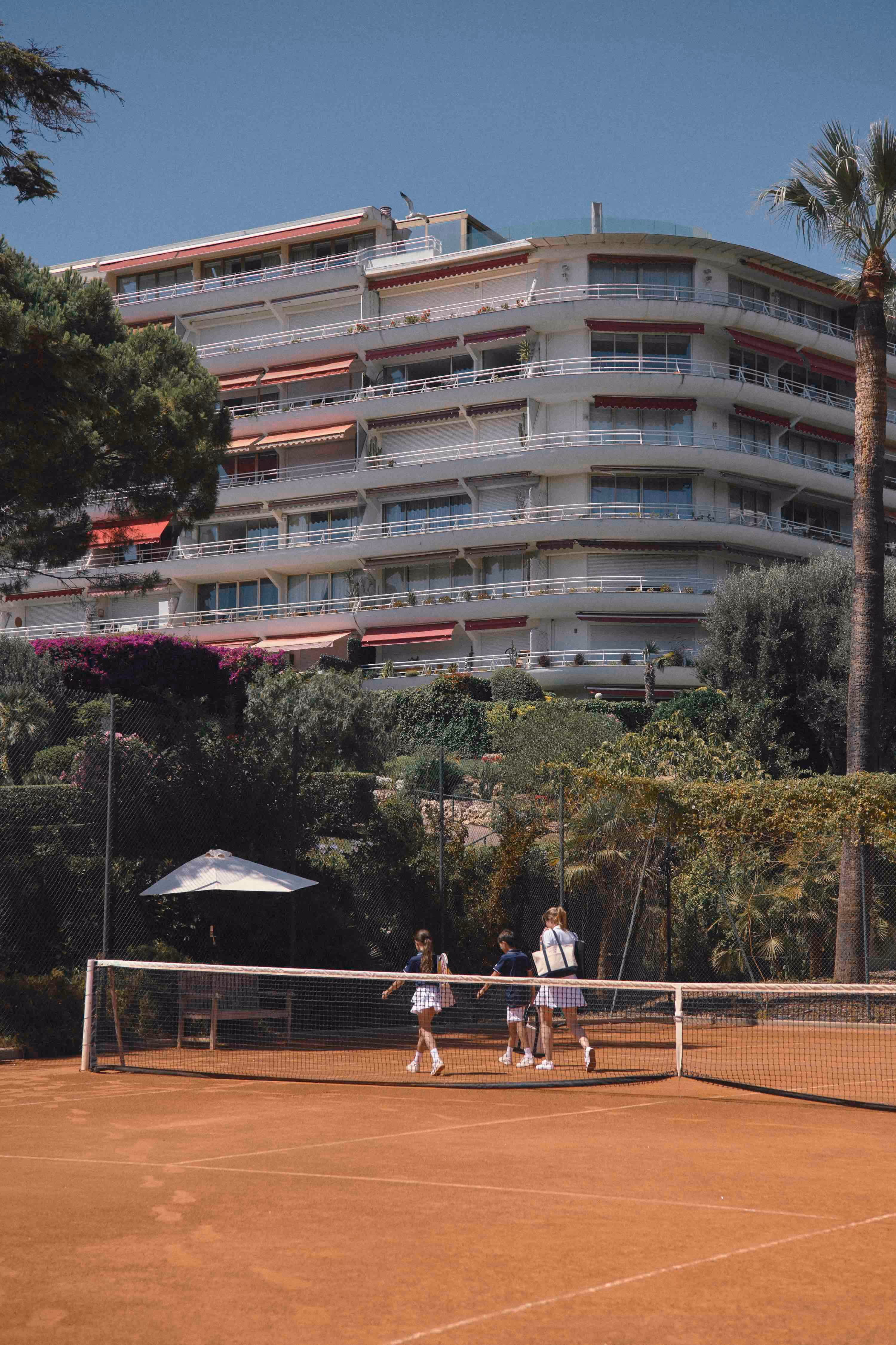 Woman and two children walking on tennis court in front of apartment blocks with blue skies and surrounding palm trees