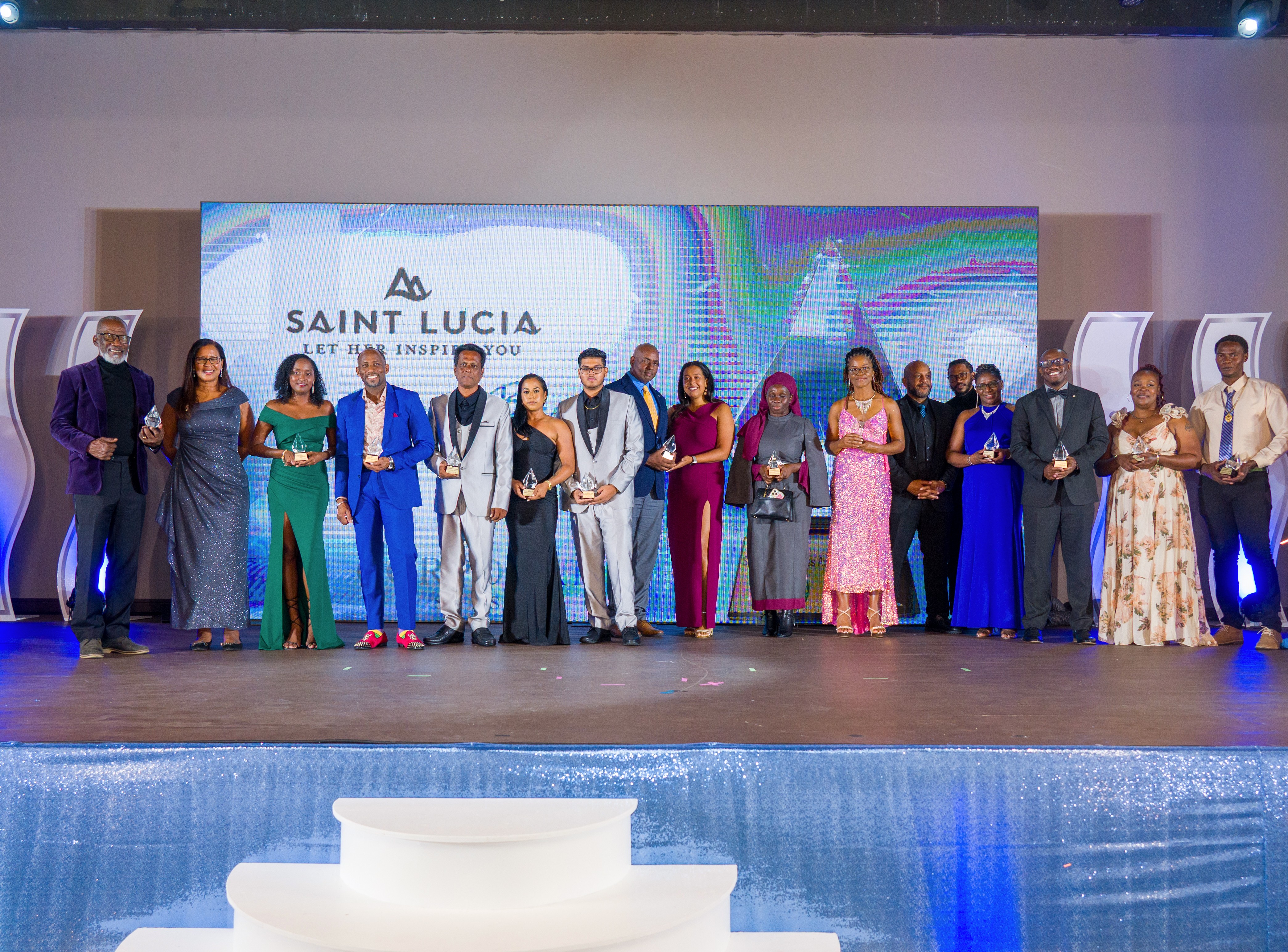 Three people standing together on stage, with a woman in the center holding a crystal award and the man on the right smiling and presenting it.