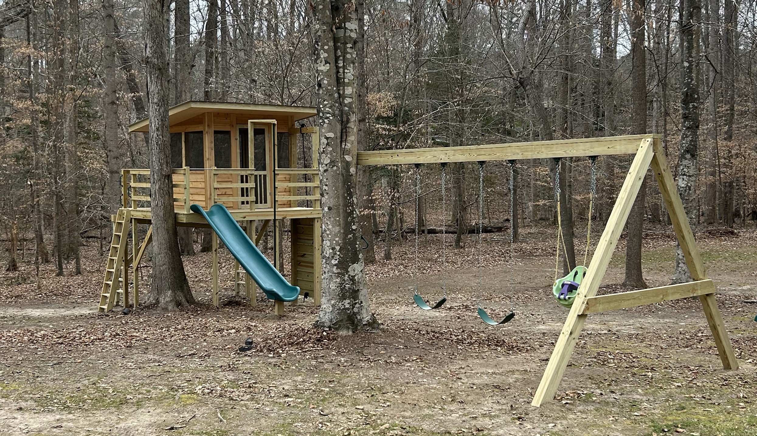 Treehouse with slide and attached set of swings. One baby swing. Set in the woods. Made of solid wood.