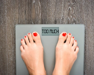 Woman staring down at her weight scale reading "TOO MUCH" signifying that she's unsure how to calculate if she is obese or not.