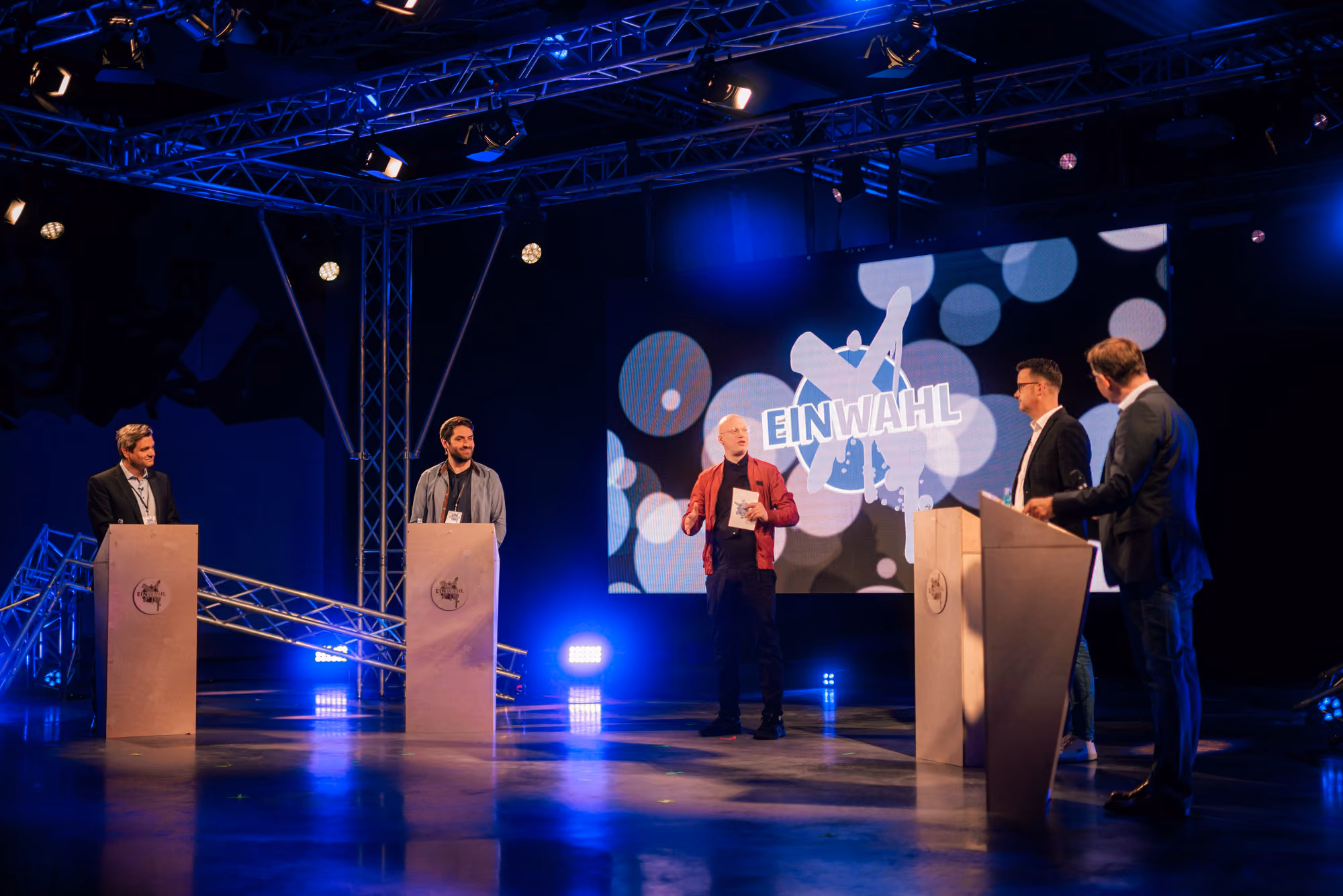 A studio setting featuring four politicians and a moderator infront of a screen displaying the EinWahl logo.