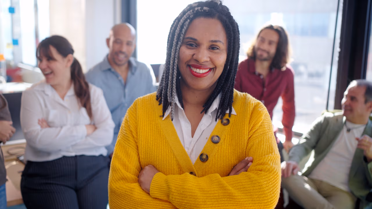 Confident businesswoman smiling in office with team members chatting behind her.