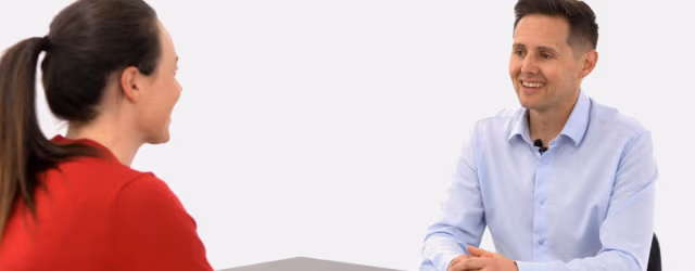 A smiling male instructor in a light blue button-down shirt sits across from a woman in a red top, illustrating a positive coaching interaction.