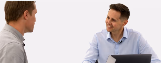 A professional male coach in a light blue shirt with a lapel microphone sits at a desk with a laptop, smiling while observing and speaking with a male team member.