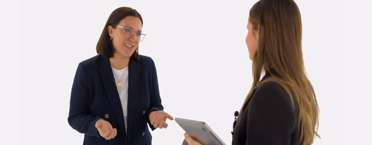A professional woman in a navy blazer smiles and gestures with her hands while providing feedback to a colleague holding a tablet.