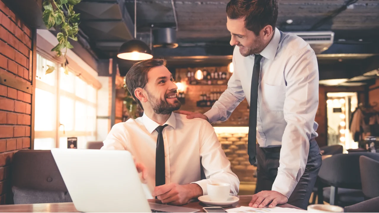 Two business colleagues in a café-style office setting collaborate over a laptop, with one standing and offering supportive guidance while the other sits smiling, suggesting mentorship, teamwork, and positive leadership.