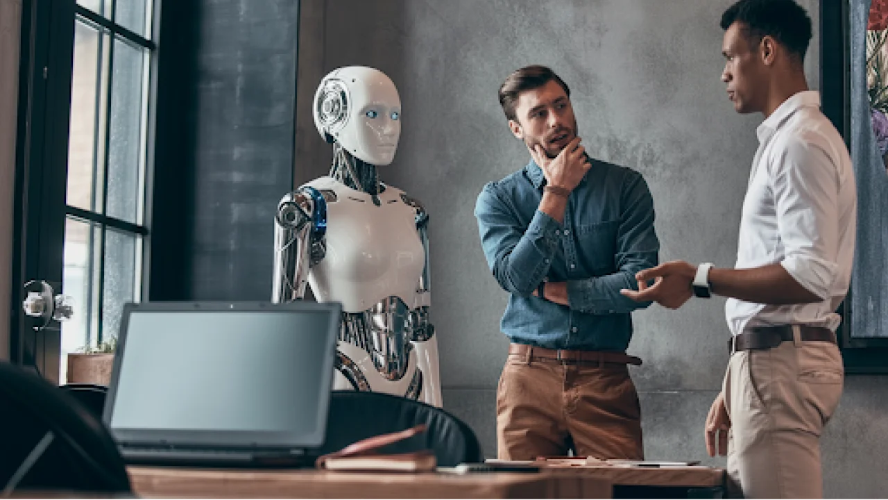 Two professionals discuss an idea in an office while a humanoid robot stands beside them, illustrating the contrast between human emotional intelligence and emerging artificial intelligence in the workplace.