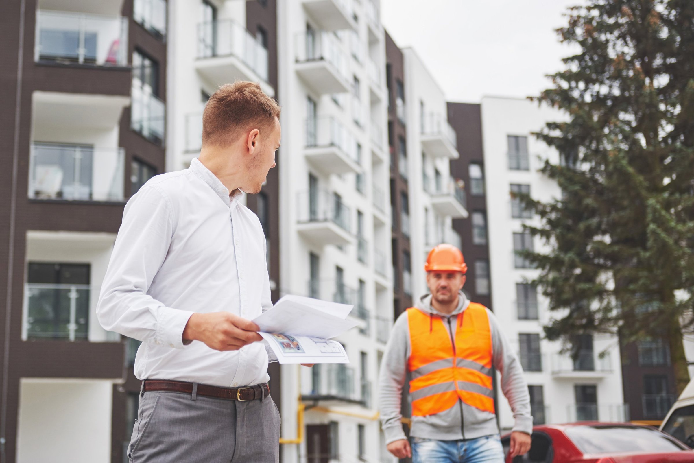 Property manager holding project documents as a contractor in a hard hat and safety vest approaches at an apartment complex