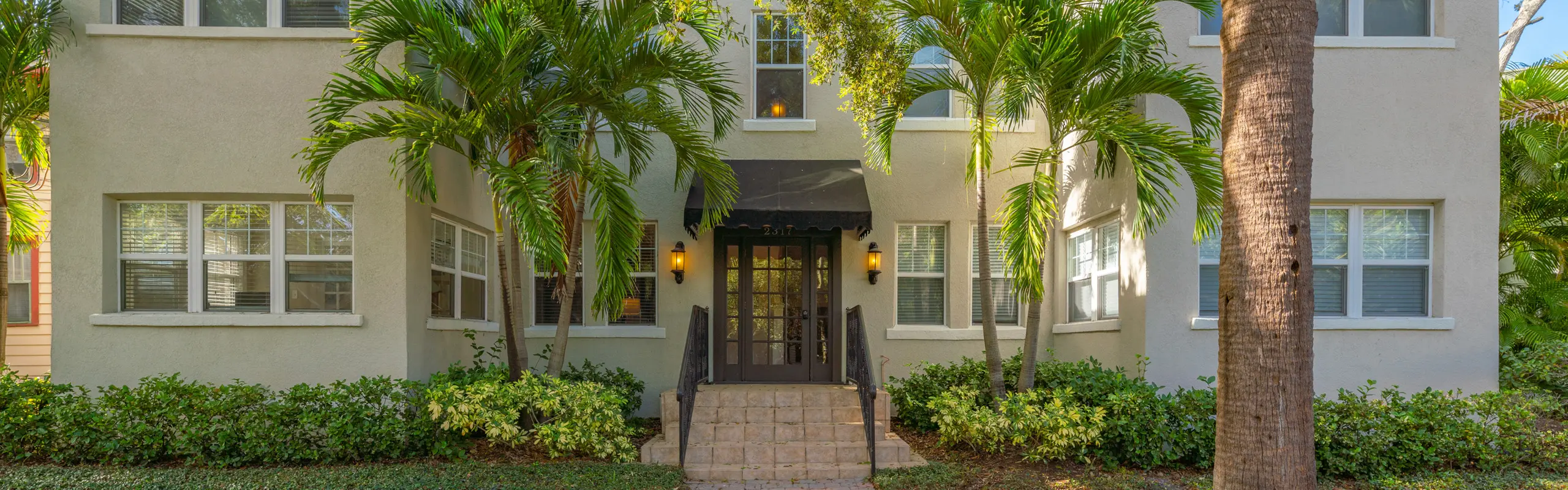Front view of an apartment complex featuring a cream exterior, palm trees, a black awning over the entryway, and lush greenery surrounding the brick path.