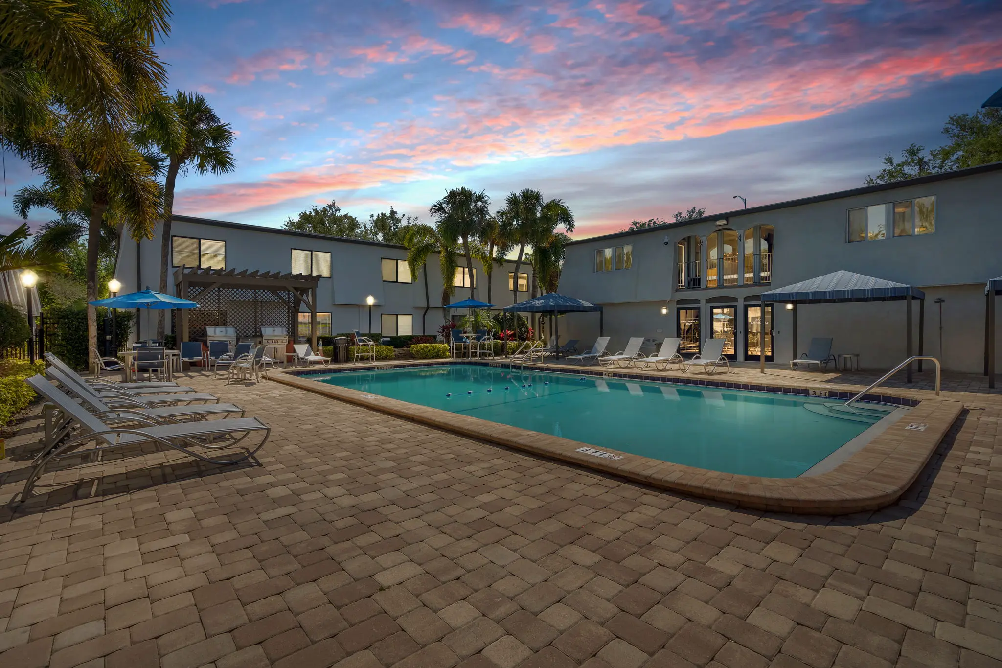 Swimming pool surrounded by lounge chairs, BBQ grills, and a white apartment complex at twilight.