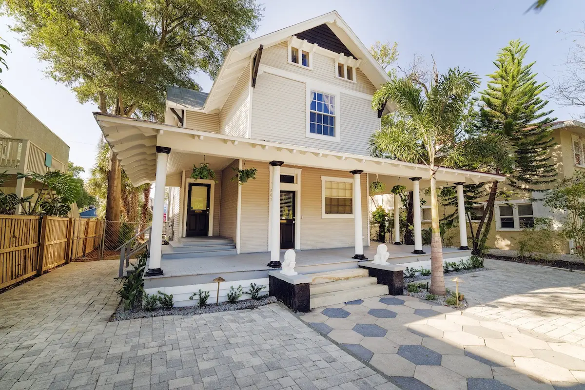 A two-story house with a white porch, black doors, and potted plants. A paved pathway and greenery surround the home.