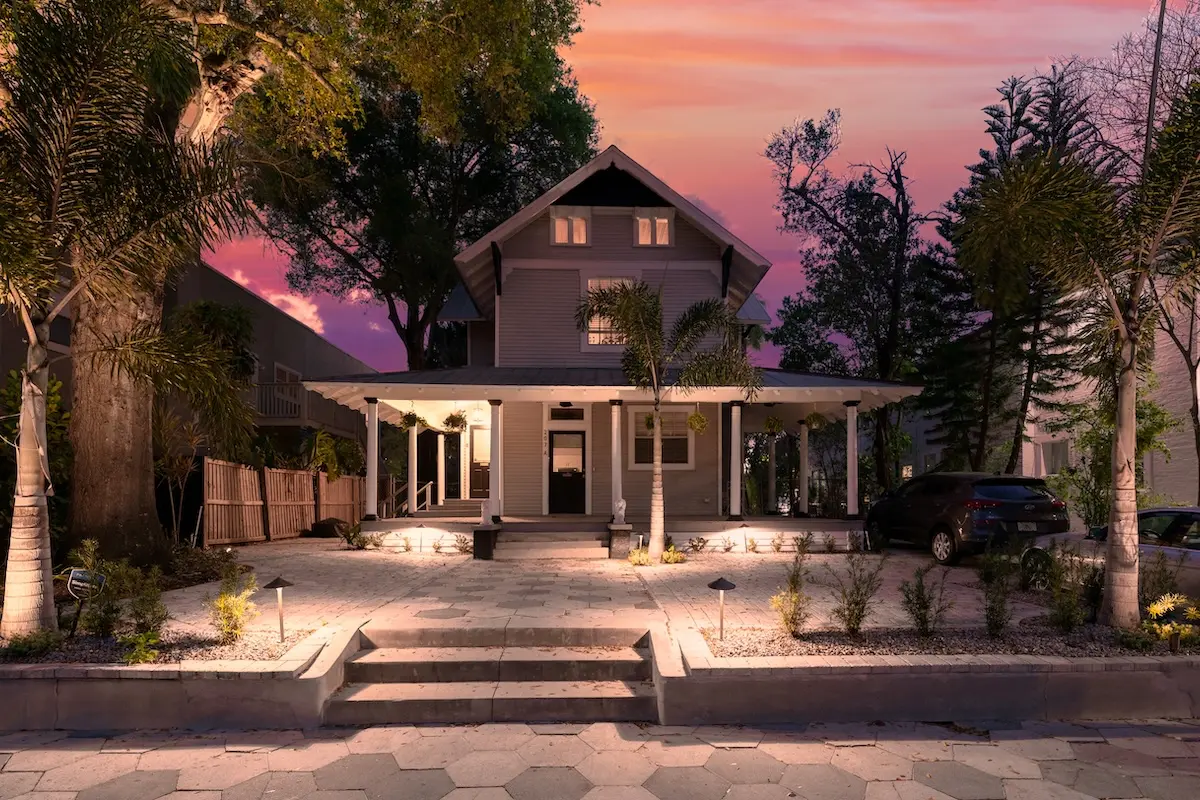 Two-story gray house at twilight with a wraparound porch, palm trees, and soft landscape lighting.