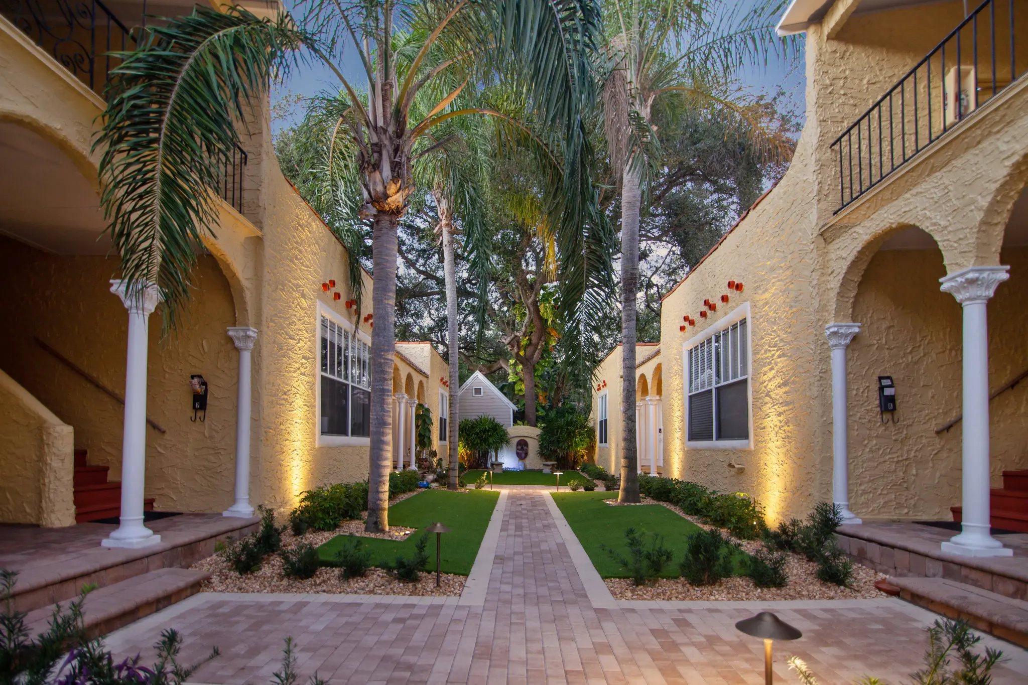 Mediterranean-style gated courtyard in Tampa’s Palma Ceia, beige stucco columns, flowering planters, and a brick walkway leading to a courtyard fountain framed by palm trees and tropical greenery.