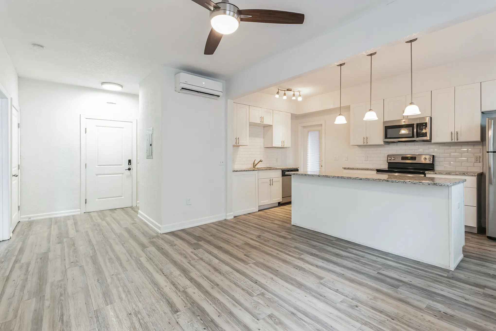 Bright, modern open-concept apartment interior with white kitchen cabinets, granite island, stainless steel appliances, pendant lighting, ceiling fan, and light wood flooring.