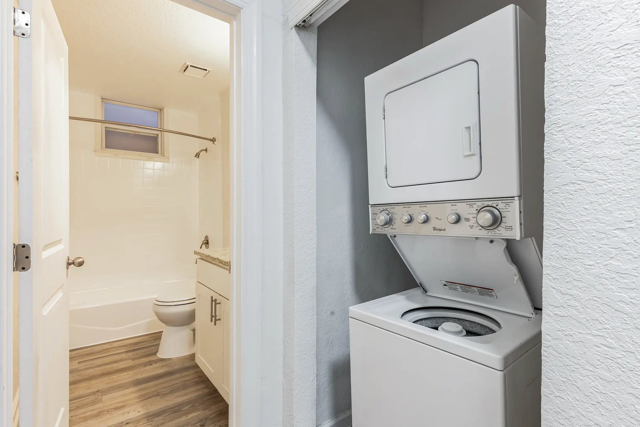 Apartment bathroom and laundry area with a stacked washer and dryer in a hallway closet next to a white bathroom featuring a bathtub, toilet, vanity, and wood-look flooring.
