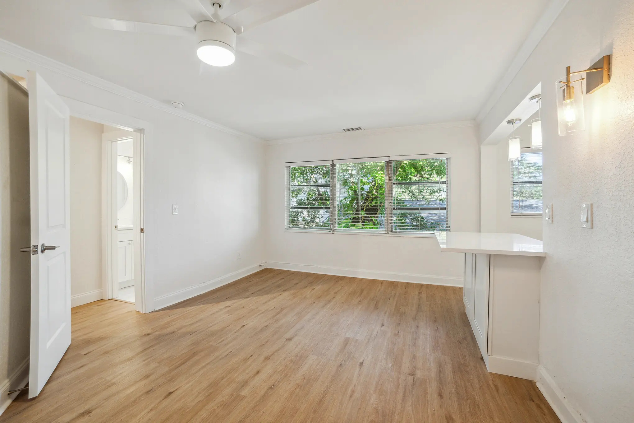 Bright, empty open-plan living area with white walls, light wood flooring, ceiling fan with light, large window with blinds and green trees outside, and a small white kitchen bar with pendant lighting.