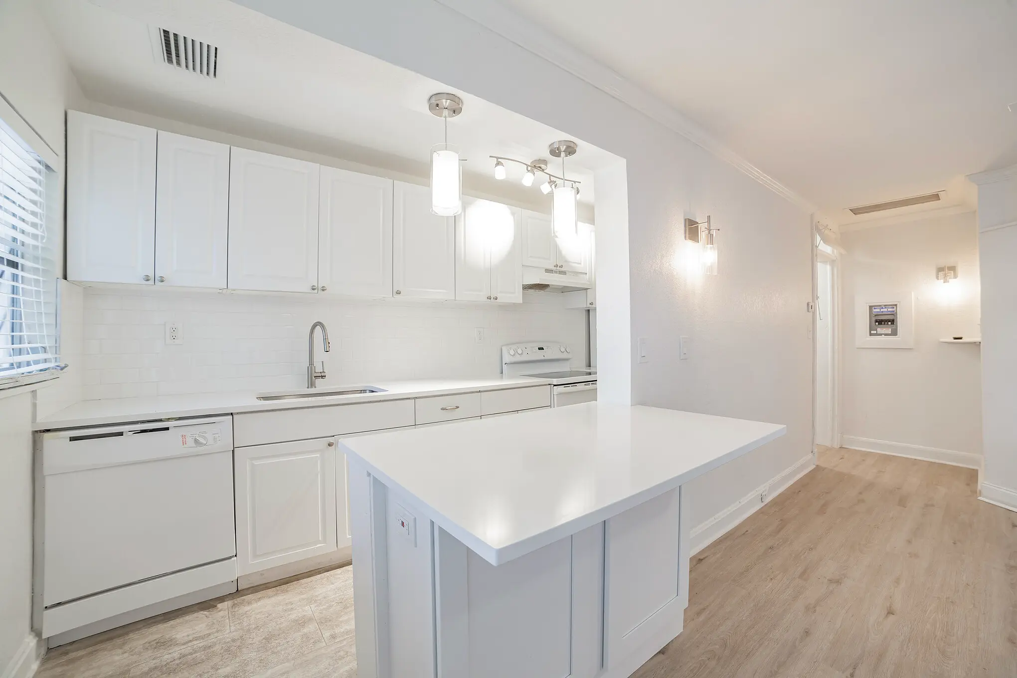 Bright white kitchen with shaker cabinets, large island, pendant lighting, subway tile backsplash, and white appliances, opening into a hallway with light wood flooring.