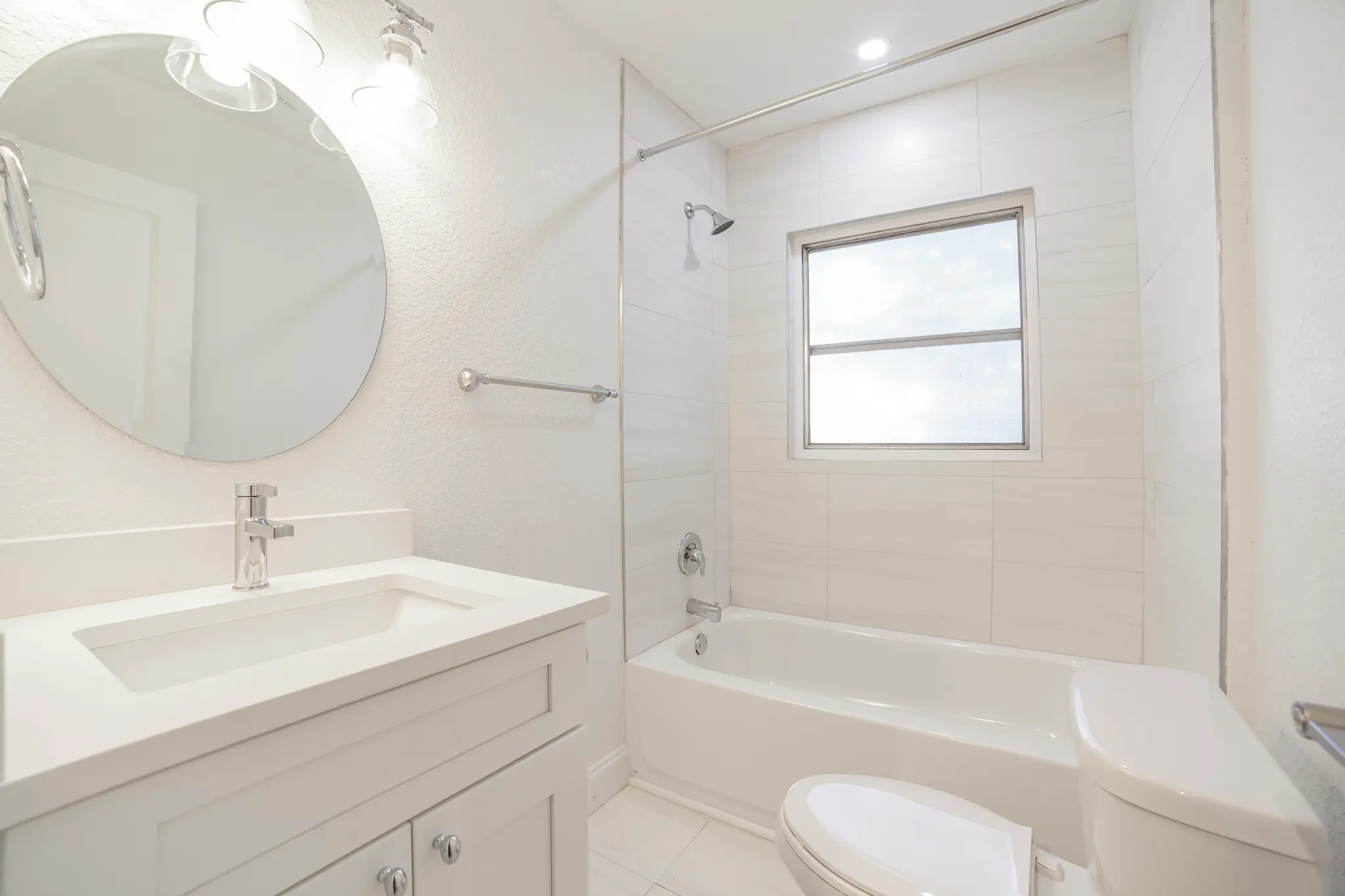 Bright white modern bathroom with a single-sink vanity, round mirror, bathtub-shower combo with glass panel, frosted window, and toilet.