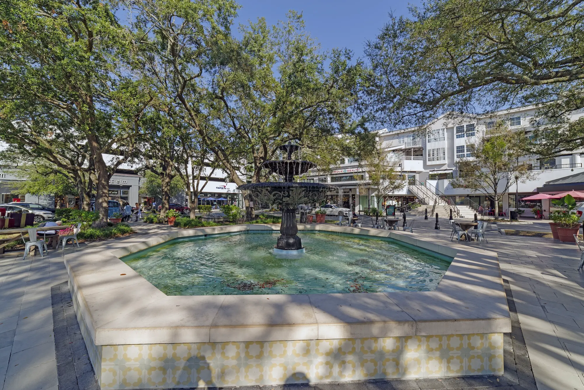 Outdoor plaza with a central tiered fountain, shaded by large oak trees, surrounded by shops, cafés, and seating on a sunny day.