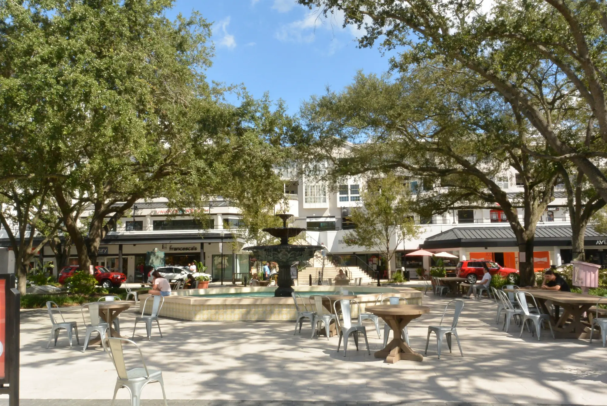 outdoor plaza with café tables and metal chairs surrounding a central fountain, lined with shops and trees on a clear day.