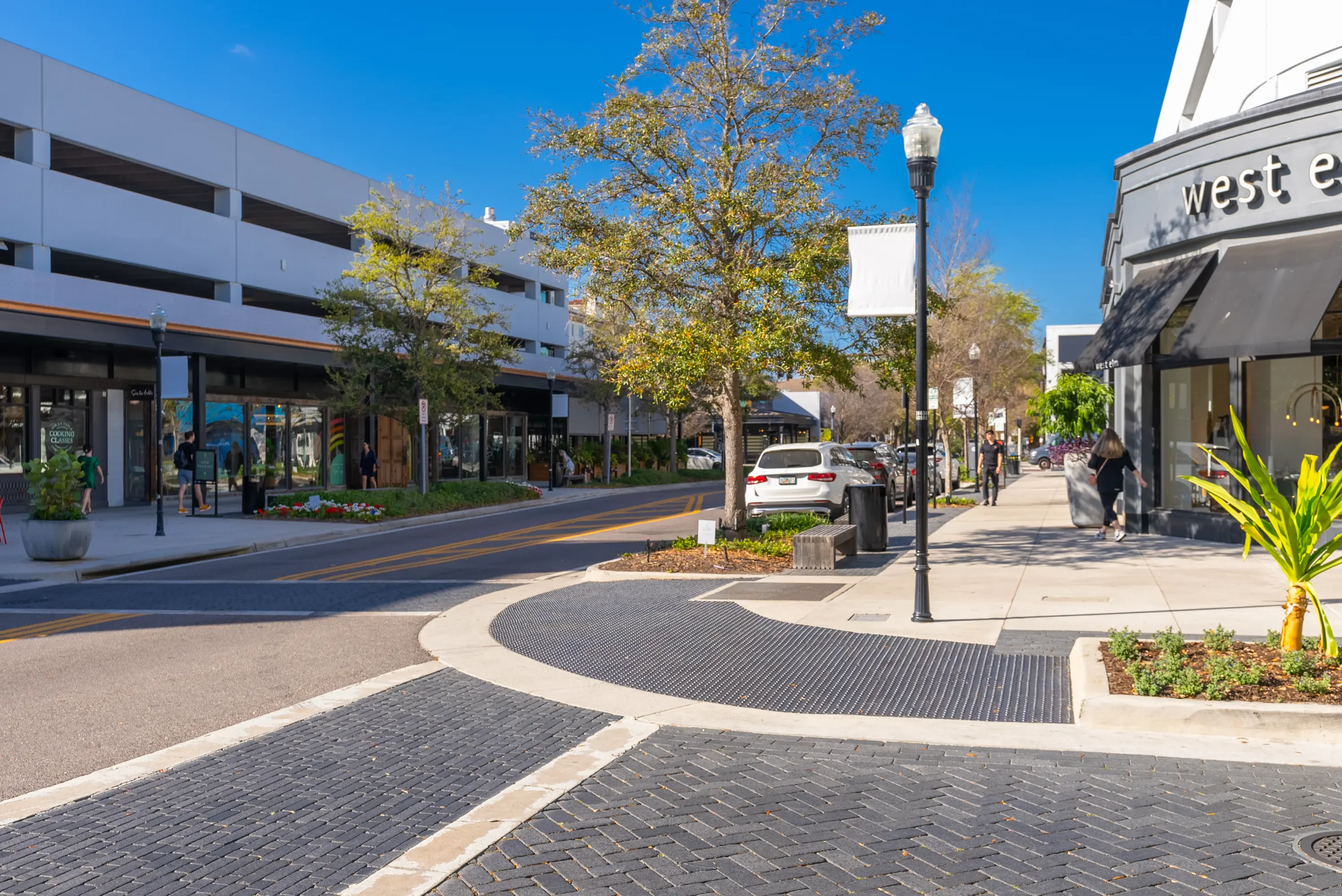 Shopping street with modern storefronts, landscaped sidewalks, parked cars, and people walking on a sunny day, featuring the West Elm store on the right and a multi-level parking garage on the left.