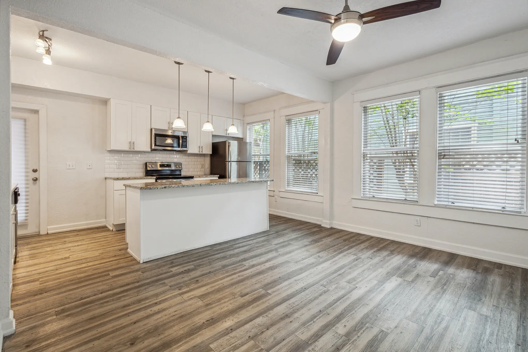 Bright, modern open-concept apartment interior with white kitchen cabinets, granite island, stainless steel appliances, pendant lighting, ceiling fan, and light wood flooring.