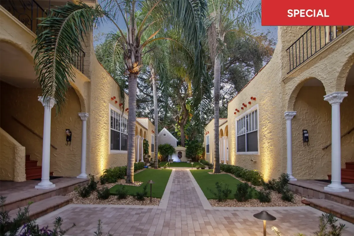 Mediterranean-style gated courtyard in Tampa’s Palma Ceia, beige stucco columns, flowering planters, and a brick walkway leading to a courtyard fountain framed by palm trees and tropical greenery with text overlay stating "special.""