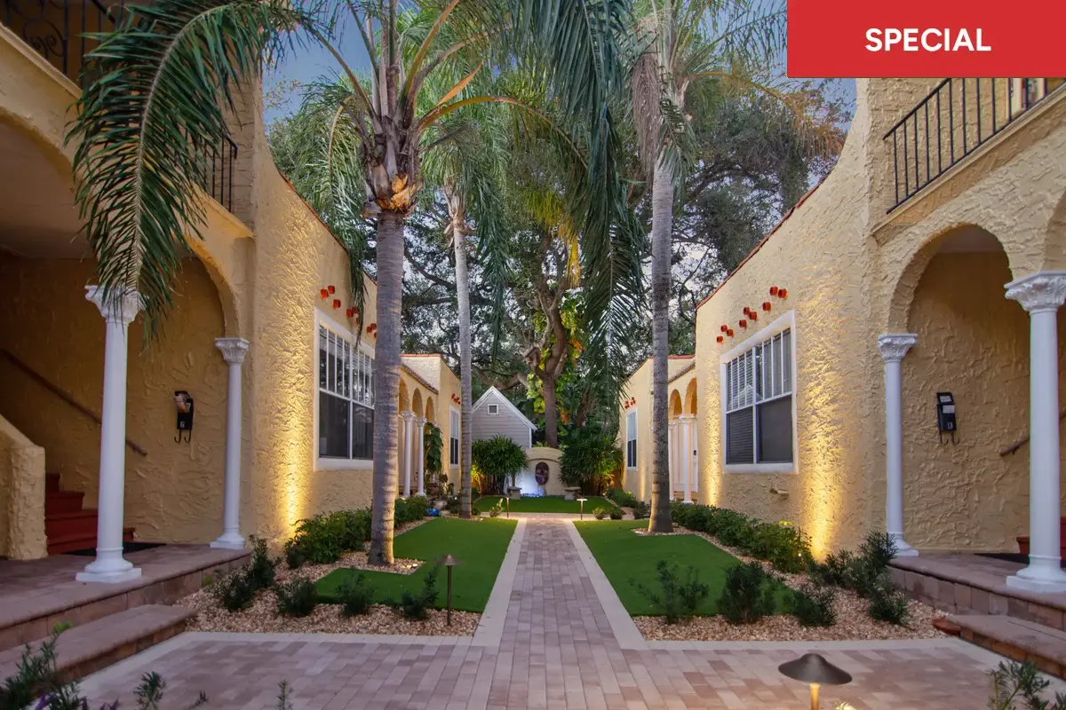 Mediterranean-style gated courtyard in Tampa’s Palma Ceia, beige stucco columns, flowering planters, and a brick walkway leading to a courtyard fountain framed by palm trees and tropical greenery with text overlay stating "SPECIAL."