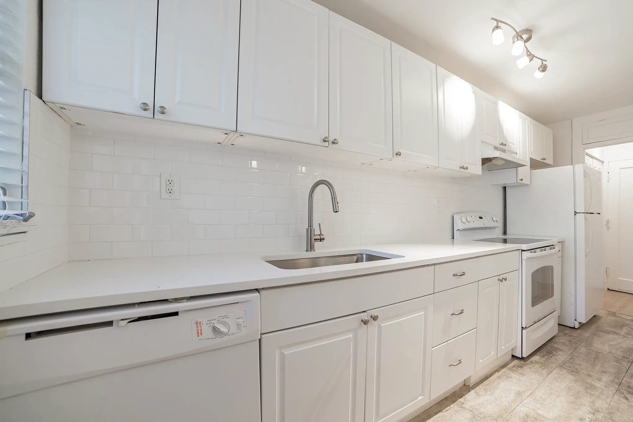 Bright white kitchen with shaker cabinets, large island, pendant lighting, subway tile backsplash, and white appliances, opening into a hallway with light wood flooring.